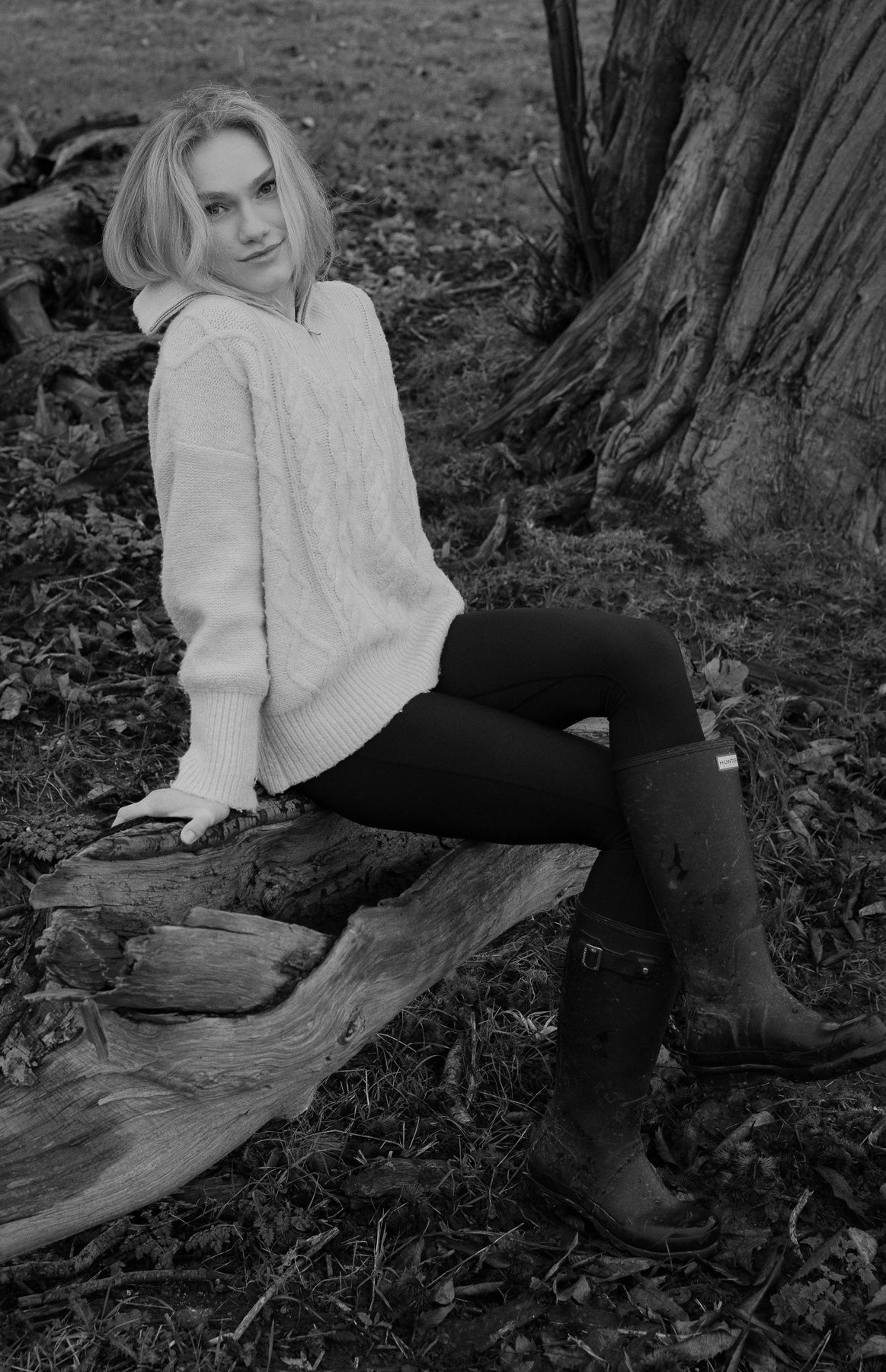 Person sitting on a log in a natural setting wearing a white sweater and black thermal leggings.
