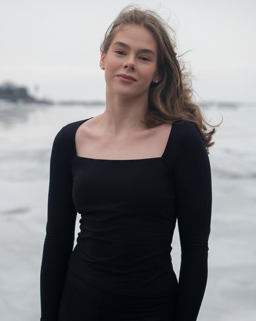 A woman wearing a thermal long sleeve black top with a square neckline from the brand Skaet, standing in front of a body of frozen water.