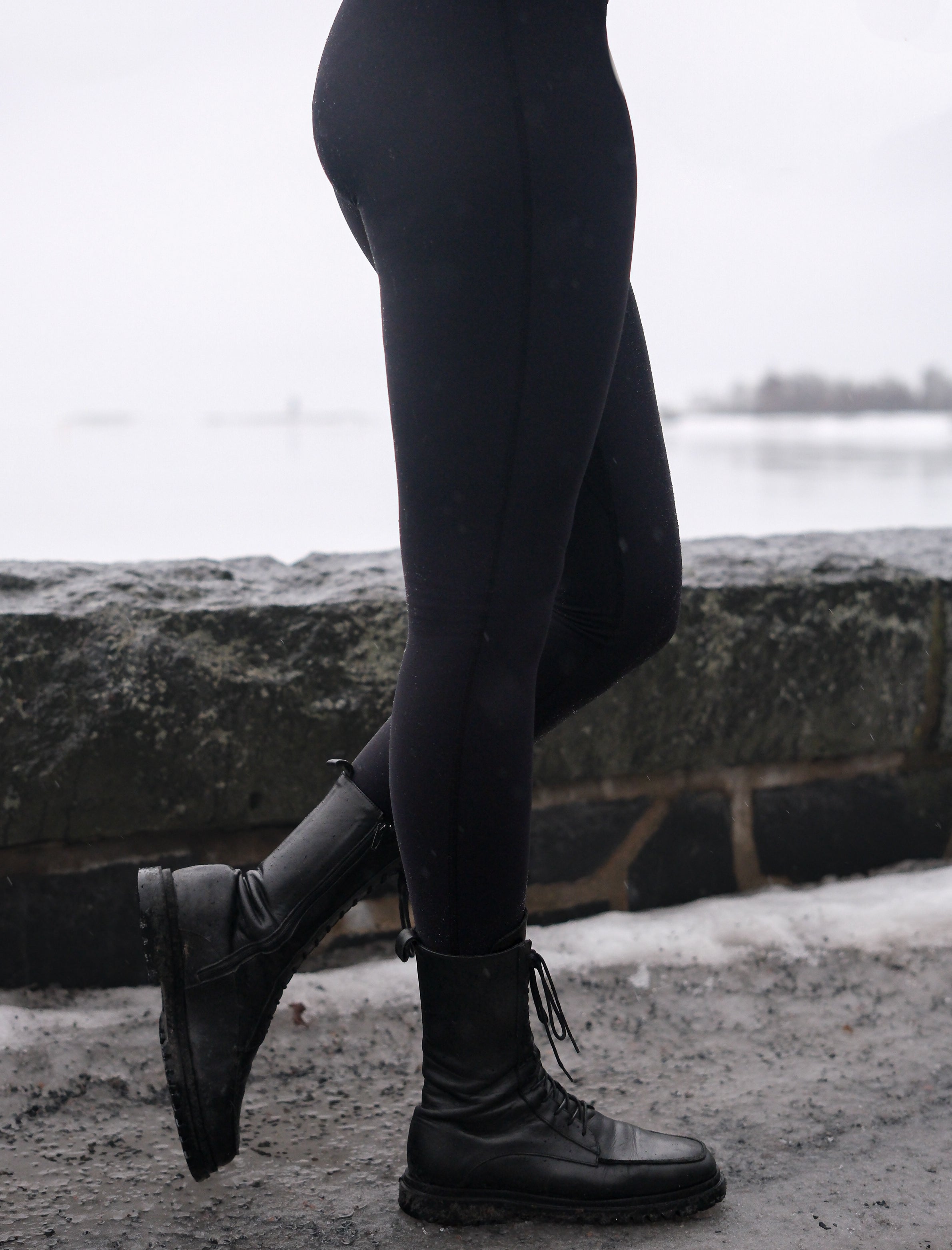 A person wearing black fleece lined leggings and black boots, standing near a body of water and a stone wall.