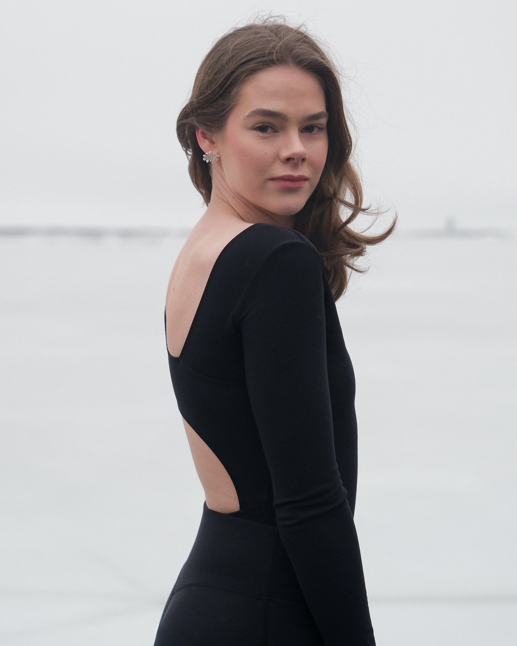 A woman looking a the camera wearing a black thermal bodysuit with asymmetrical cutouts on the back from the brand Skaet
