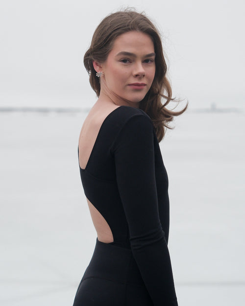 A woman looking a the camera wearing a black thermal bodysuit with asymmetrical cutouts on the back from the brand Skaet