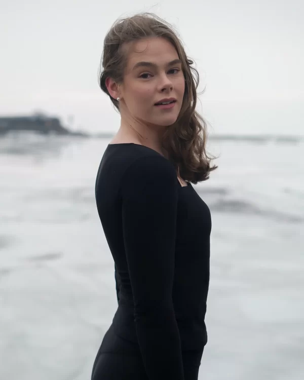 A model wearing the Skaet Long Sleeve thermal sportswear top. She is looking over her shoulder whilst near the coast with waves breaking behind her.